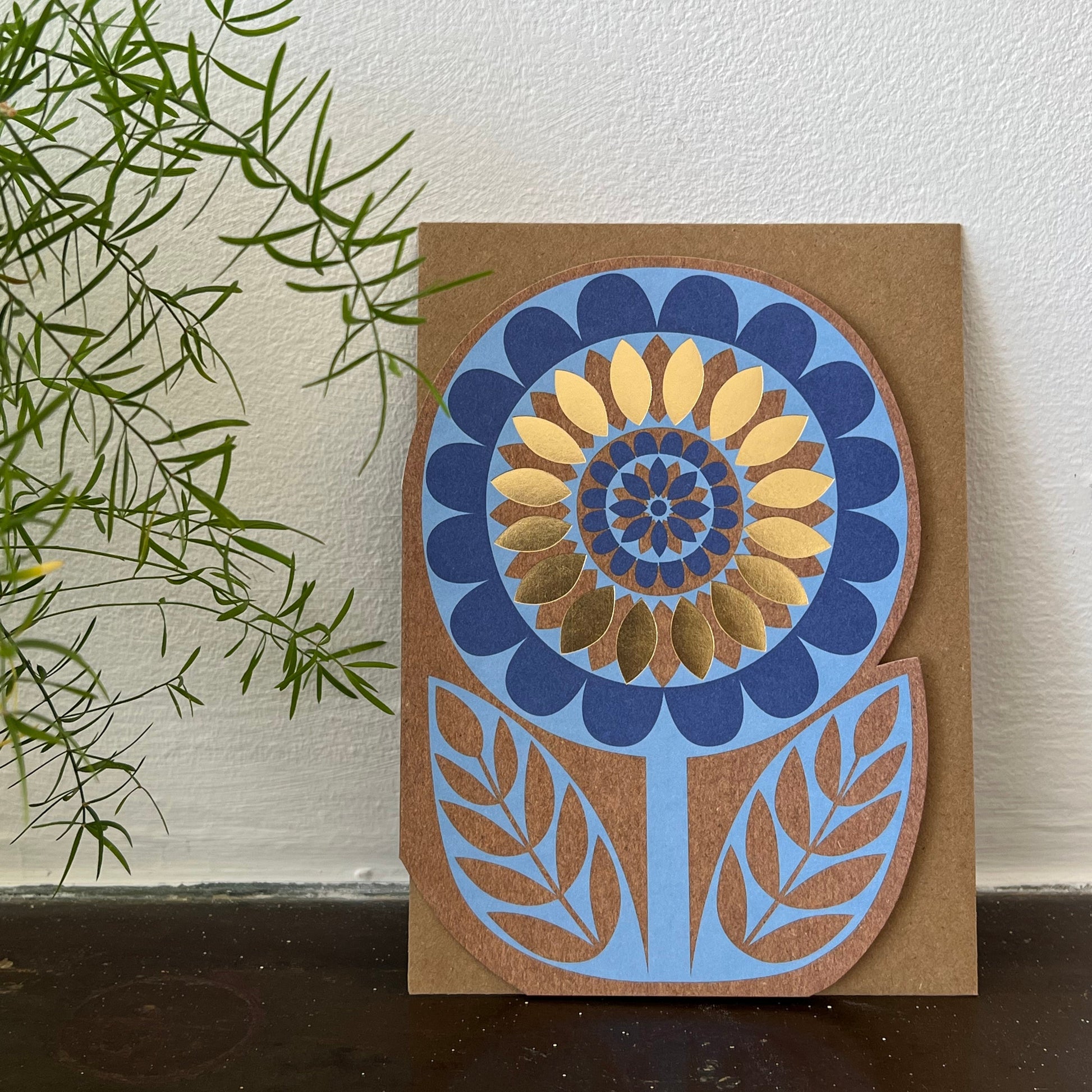 Decorative card with a blue and gold floral design on a brown background, placed on a white surface with green leaves.